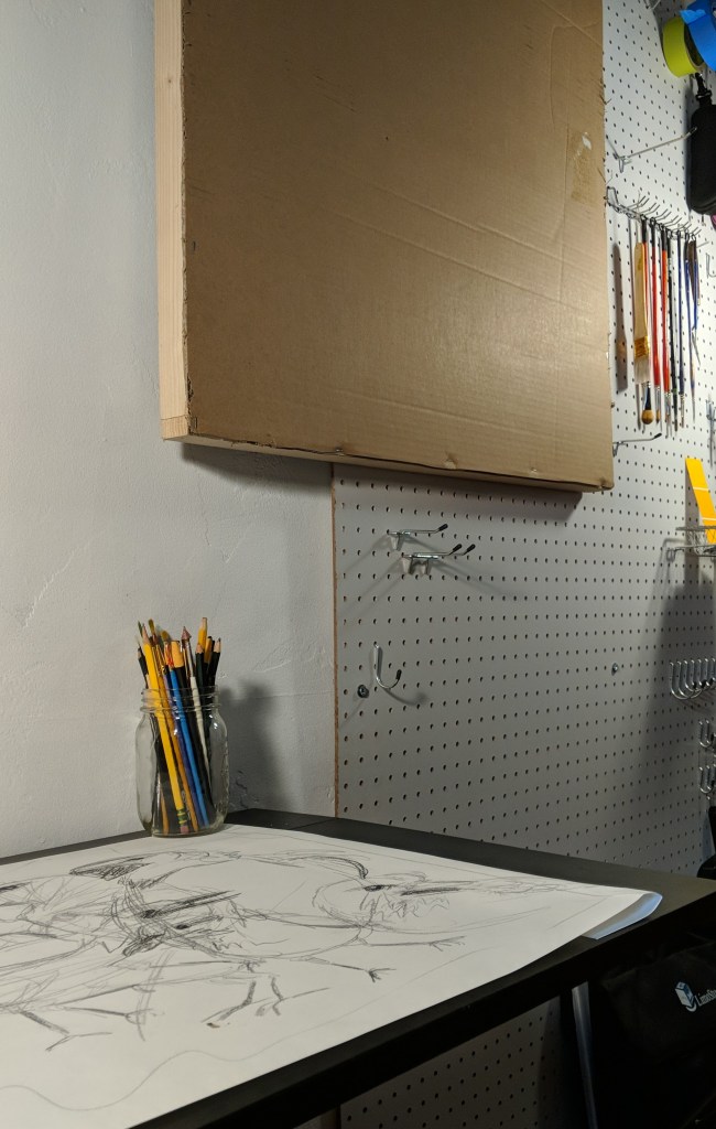 Interior close-up photo of a studio space, showing peg-board wall with hanging paint brushes and a blank art board made with a wooden frame covered in cardboard. Beside this is a small black desk with a large sketch of several birds.